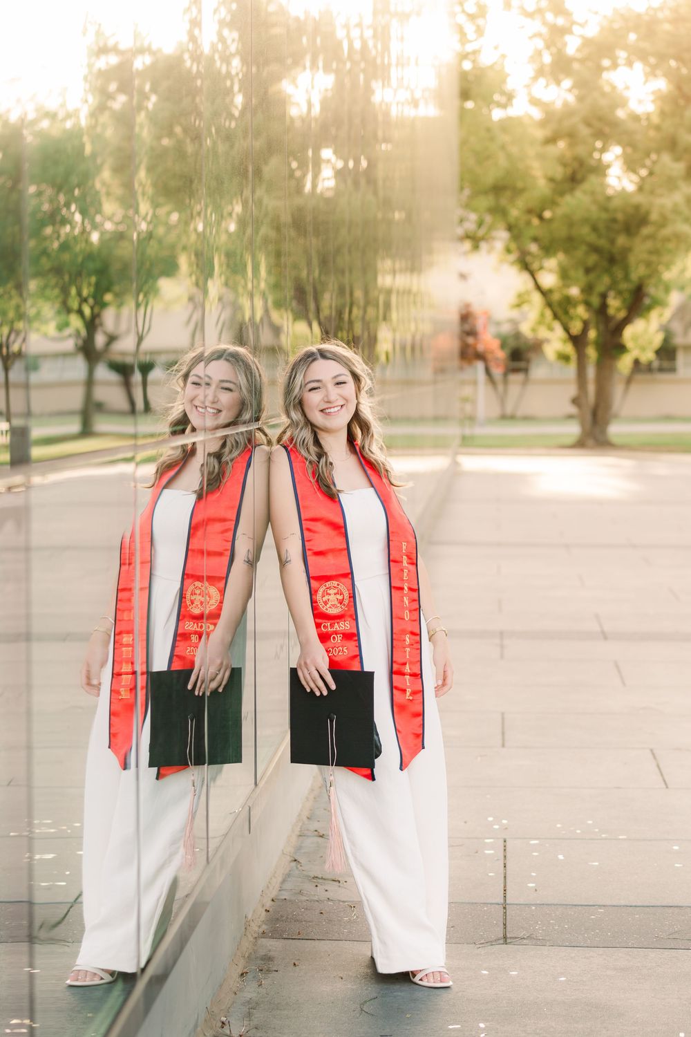 Graduate in white dress and red stole poses joyfully in front of campus building during golden hour sunset.