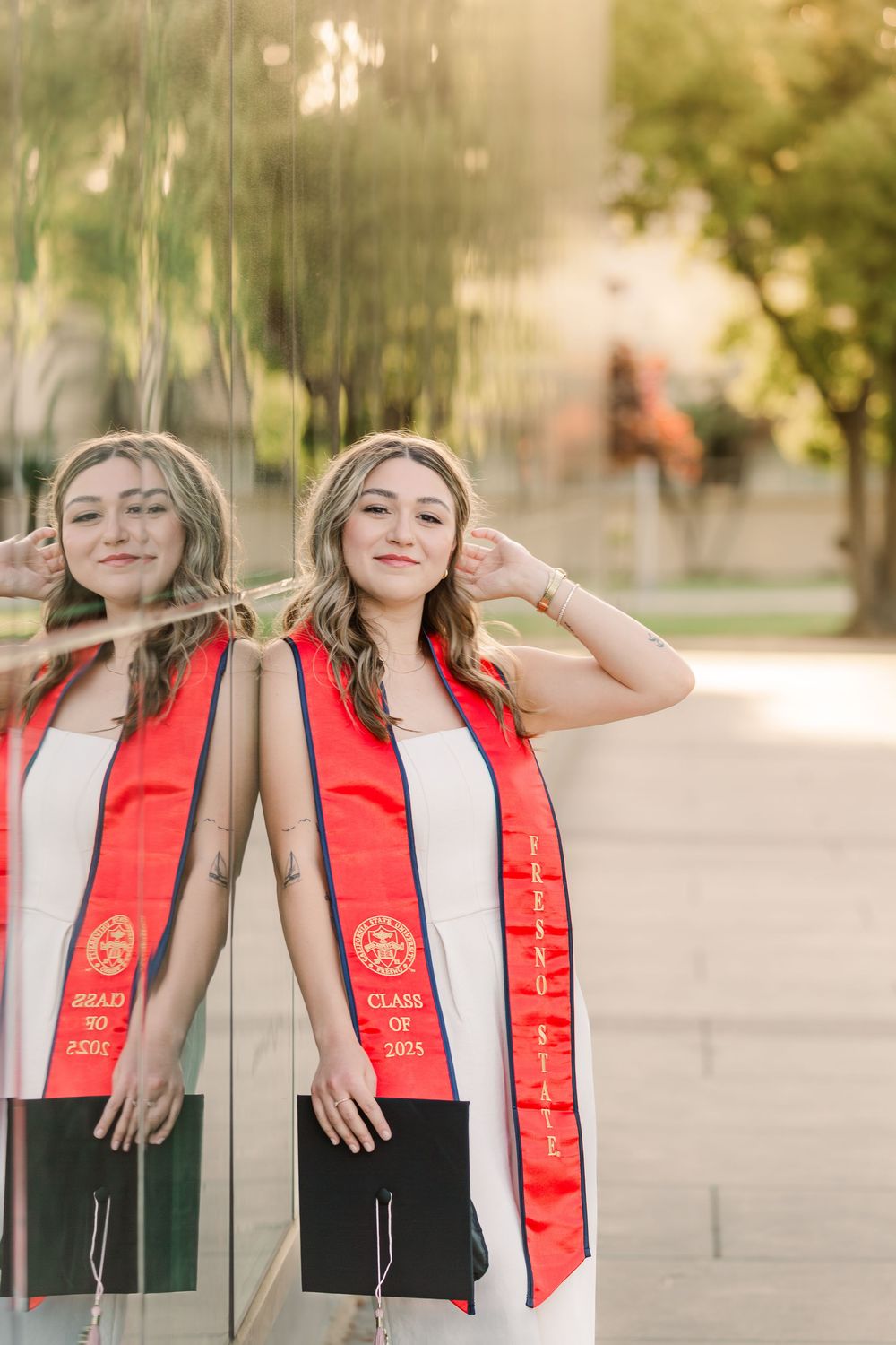 Graduate wearing red stole and white dress posing playfully during outdoor photo session.