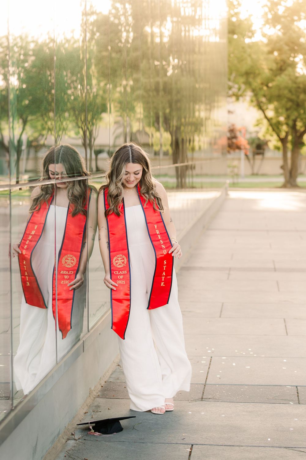 Graduate in white dress and red graduation stole poses by reflective glass wall during sunset.
