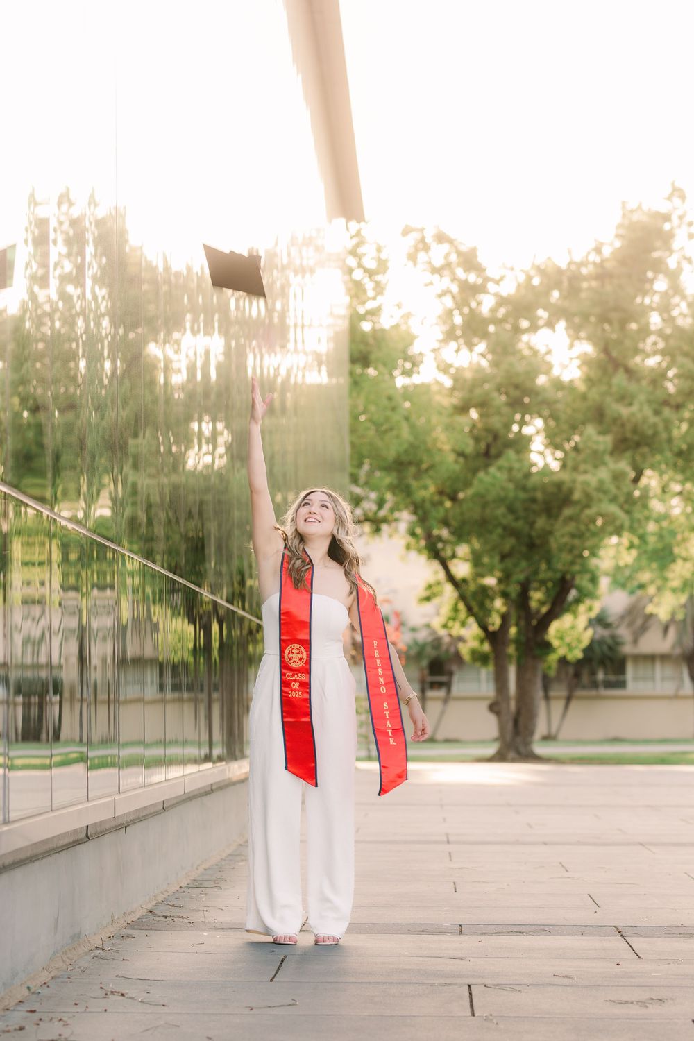 Graduate in white dress and red sash poses joyfully outdoors during sunset with trees in background.
