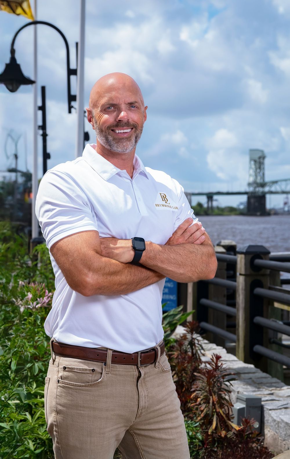 Professional wearing a white polo shirt and khaki pants poses with crossed arms on a waterfront boardwalk.
