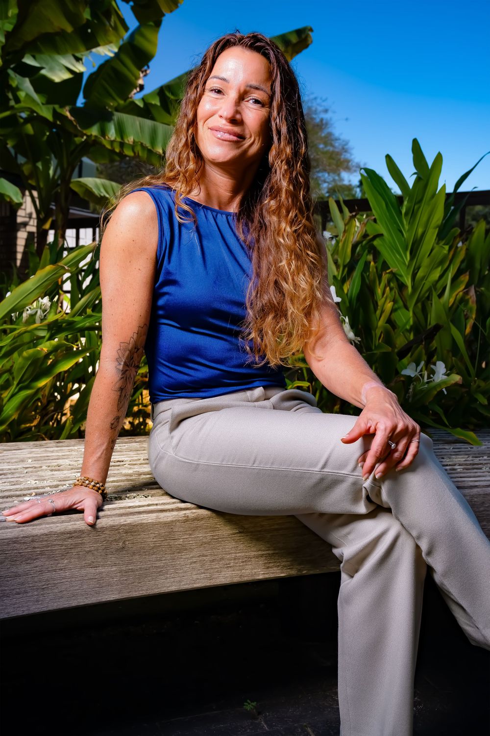 Person in blue sleeveless top and light pants sits casually on wooden bench against tropical foliage backdrop.