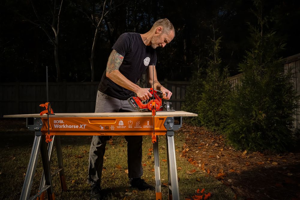 Person using a power saw to cut wood on a portable workbench in an outdoor setting.