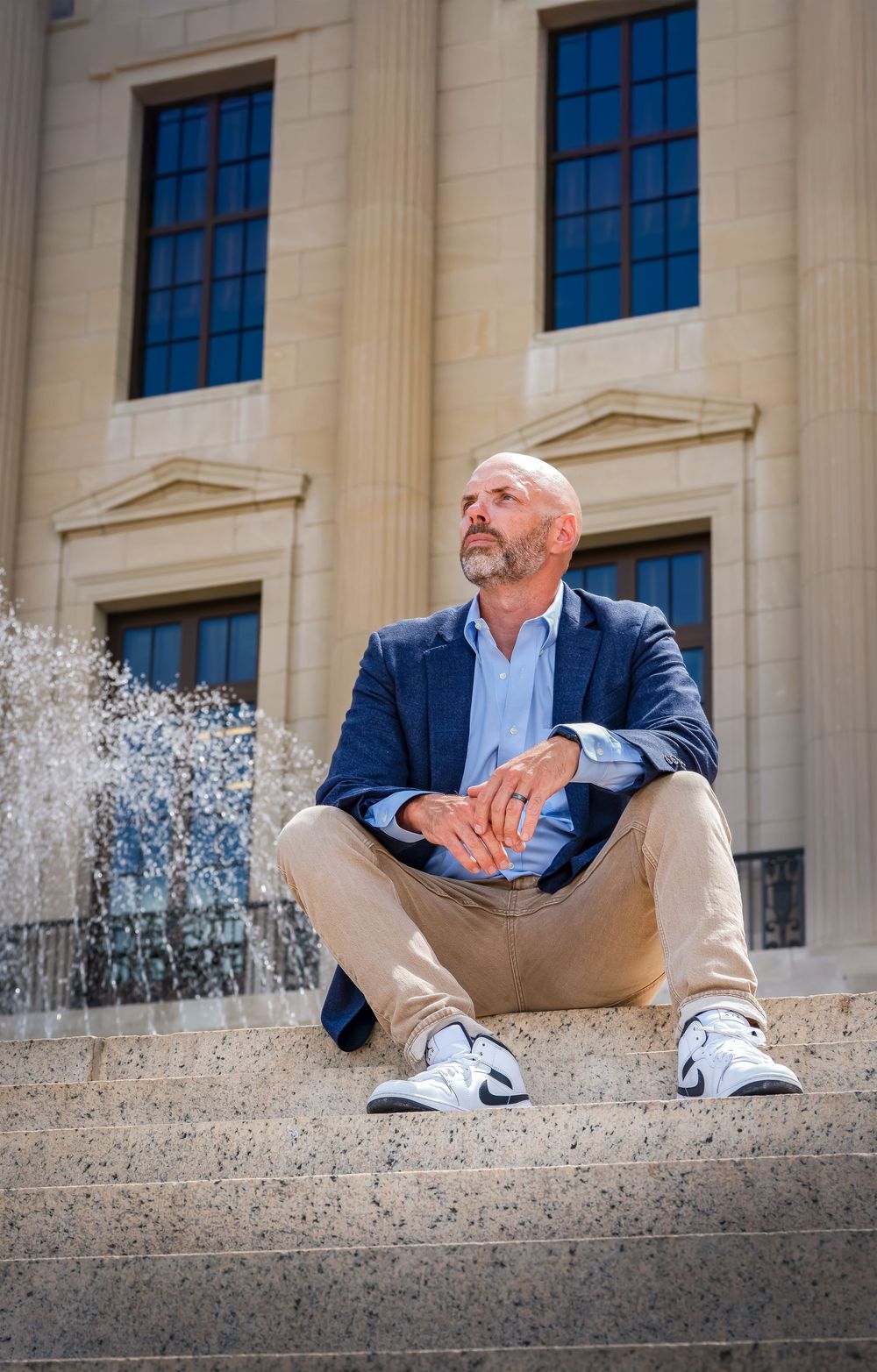 Person in blue blazer and khakis sits casually on stone steps with columns and windows in background.