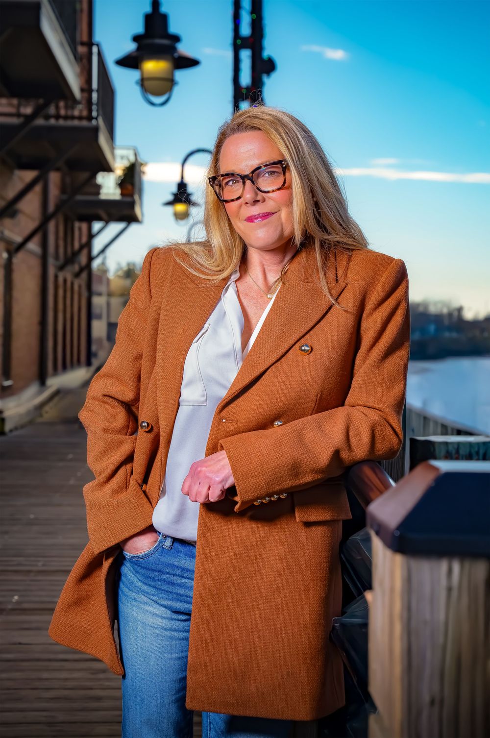 A person wearing a brown blazer, glasses, and jeans poses against a waterfront backdrop on a sunny day.