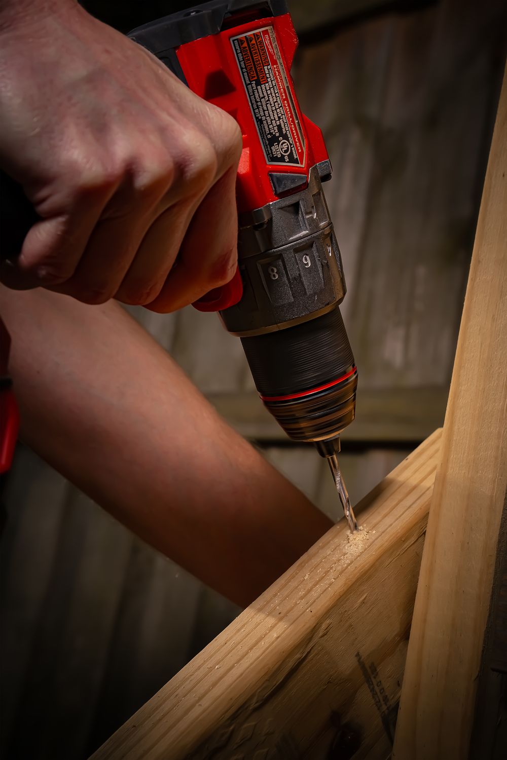 Close-up of hands using a red cordless drill to drive a screw into wooden lumber.