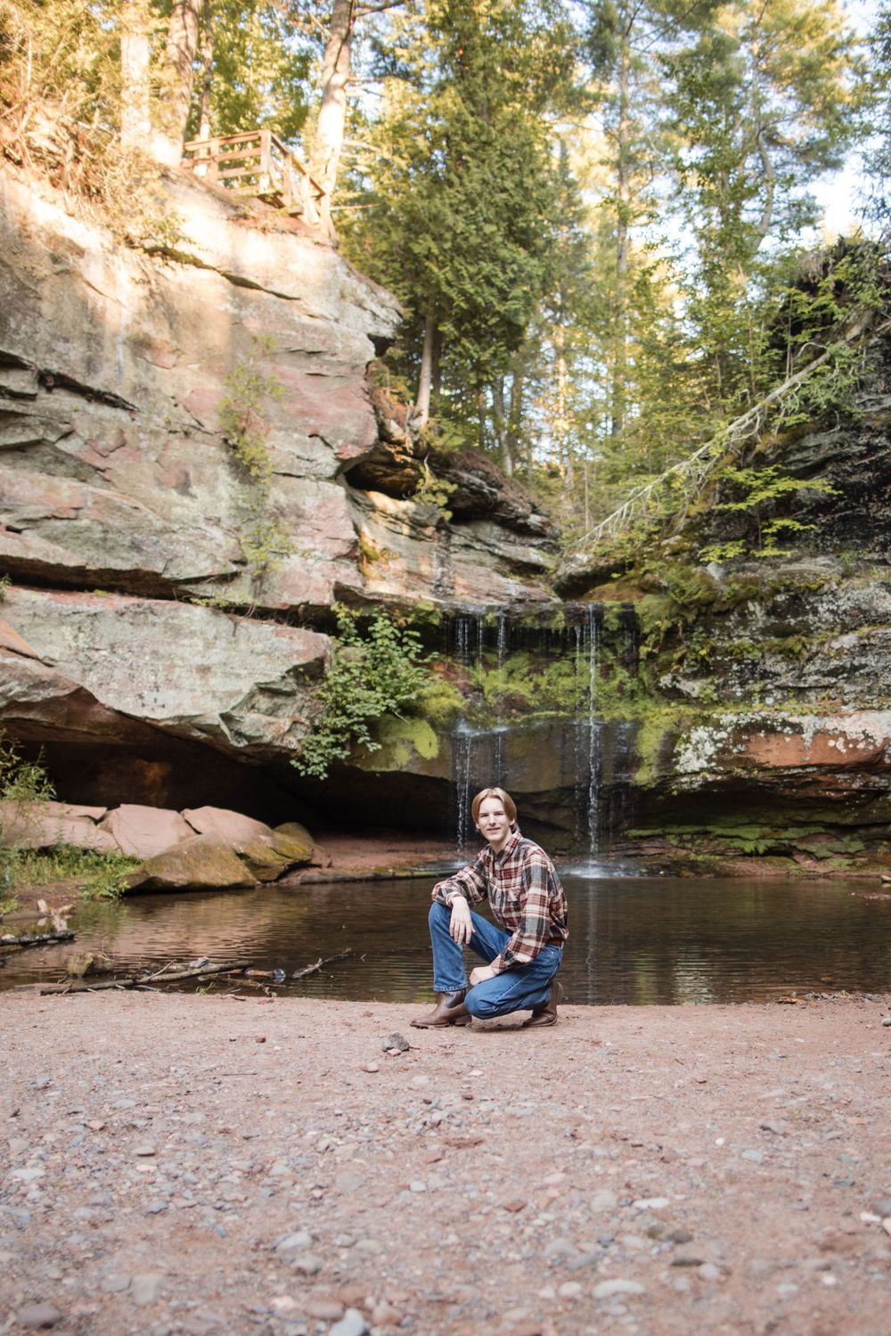 A scenic waterfall cascades down layered rock formations in a tranquil forest setting with sandy ground below.