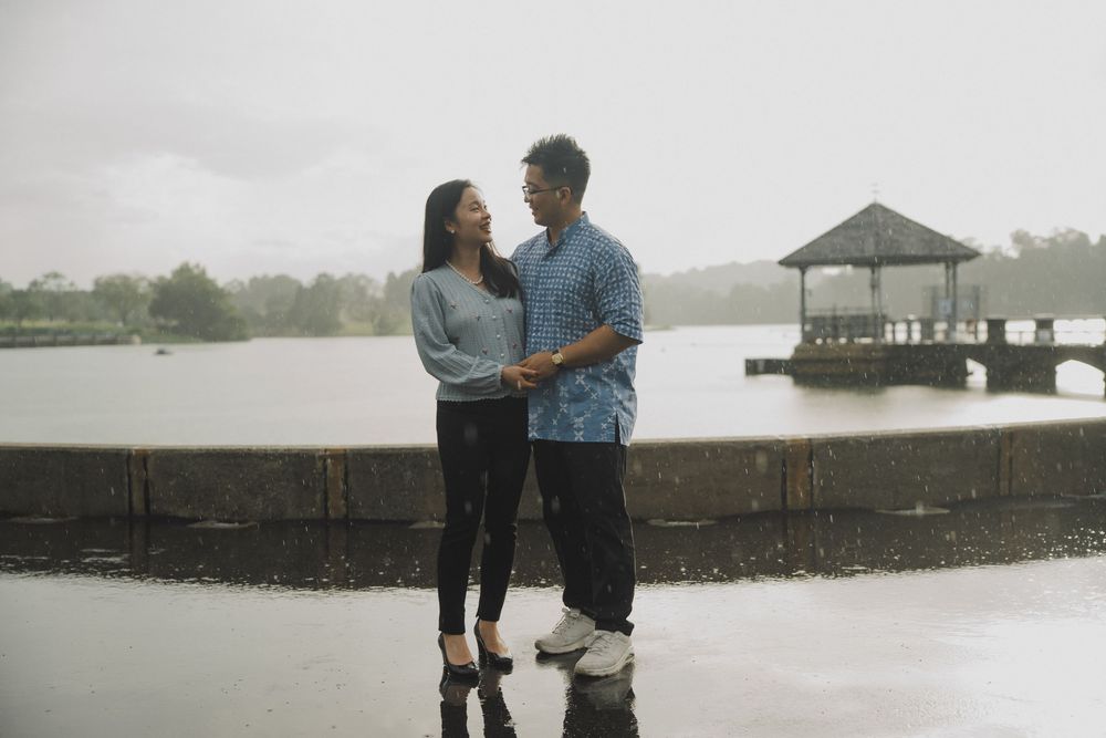 A couple shares a romantic moment on a rainy day at a lakeside pier with umbrellas and overcast skies.