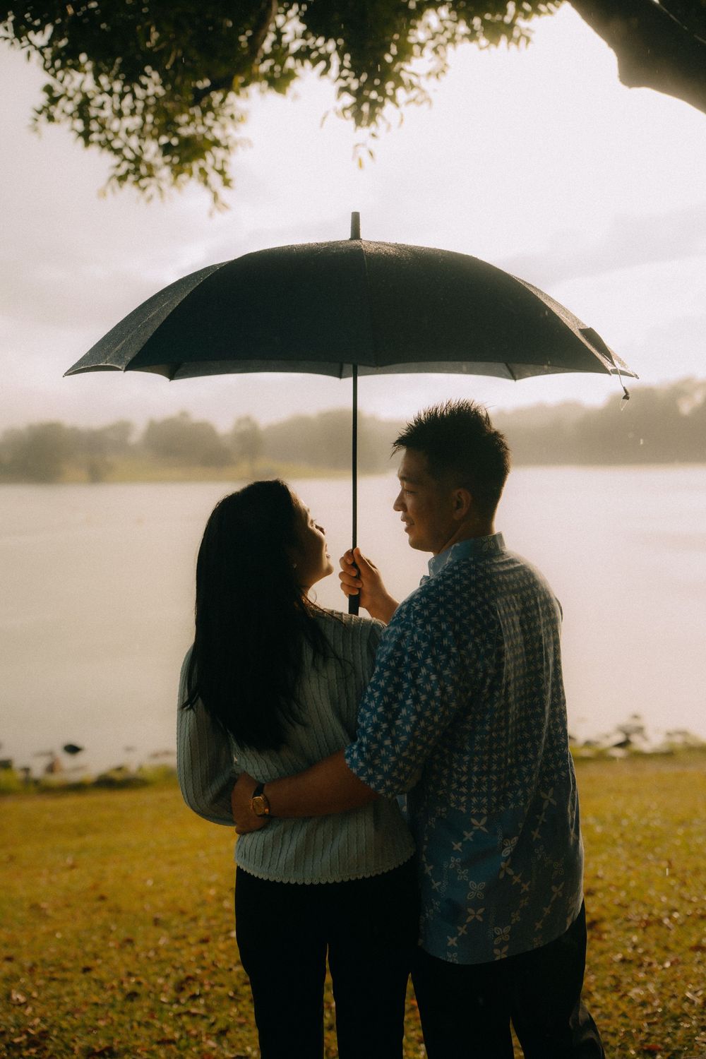 A couple shares an intimate moment under a black umbrella on a misty field by the water at sunset.