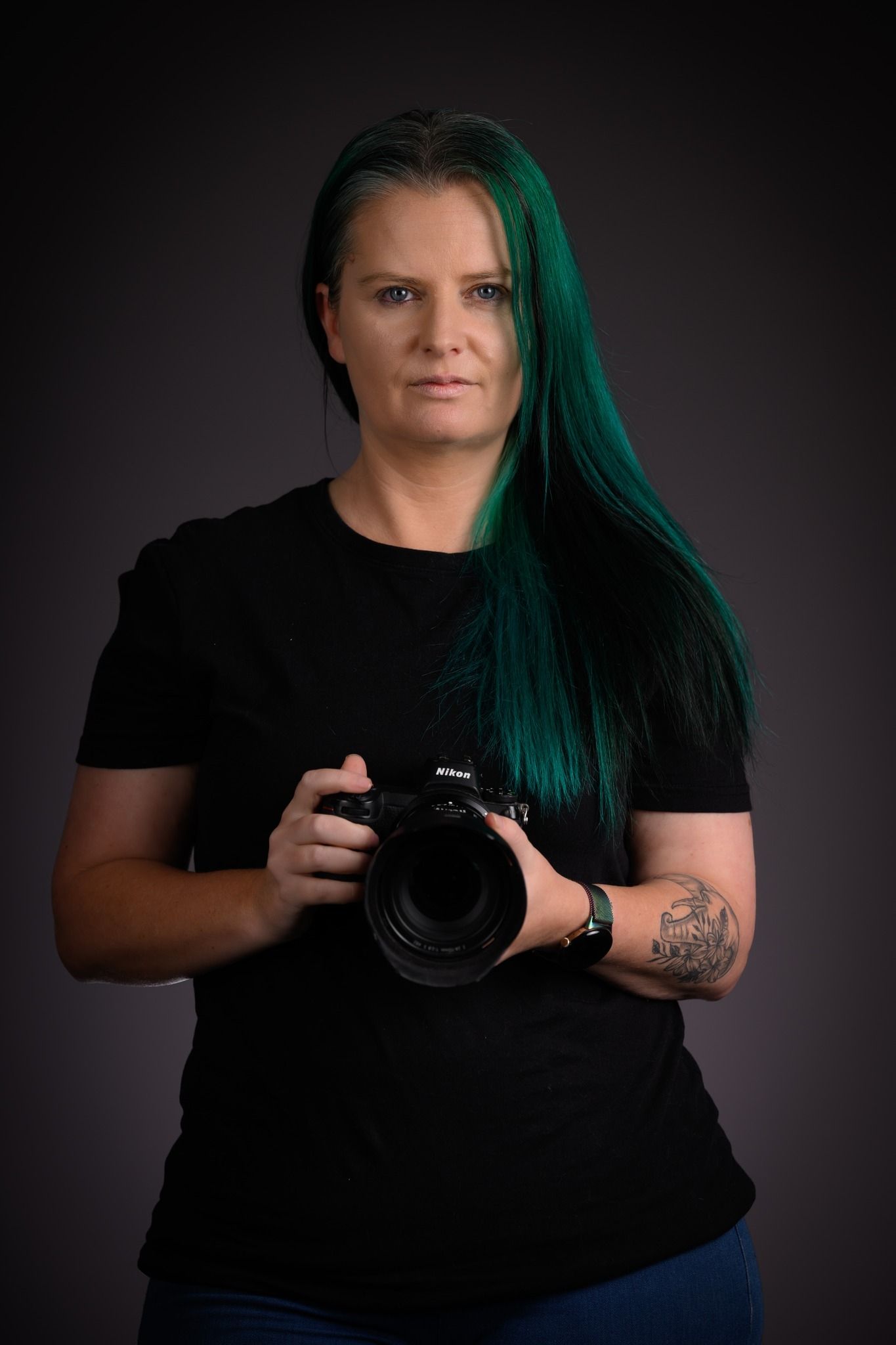 Professional photographer in black shirt holding camera against dark studio backdrop with striking teal hair.