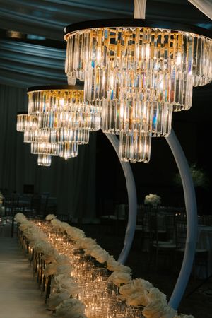 Elegant tiered crystal chandeliers illuminate a luxurious wedding reception with white floral centerpieces and candlelit table.
