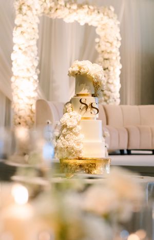 Elegant white and gold multi-tier wedding cake with floral decorations and monogram, displayed before a floral arch backdrop.