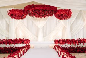 Elegant wedding aisle with red rose petal decorations, floral ceiling installation, and white draped backdrop.