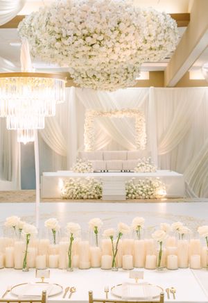 Elegant all-white wedding reception with floral chandelier, draped backdrop, candle centerpieces, and lush flower arrangements.