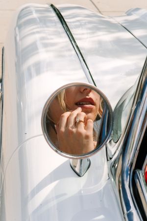 Woman applying red lipstick reflected in a vintage car side mirror, wearing a diamond ring.