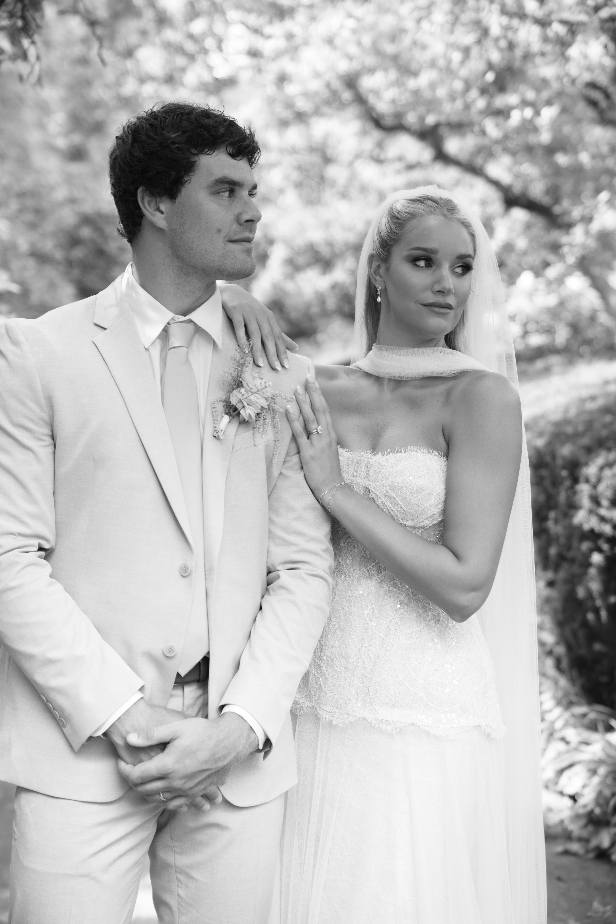 Black and white wedding photo of bride in strapless gown and groom in light suit posing outdoors among trees.