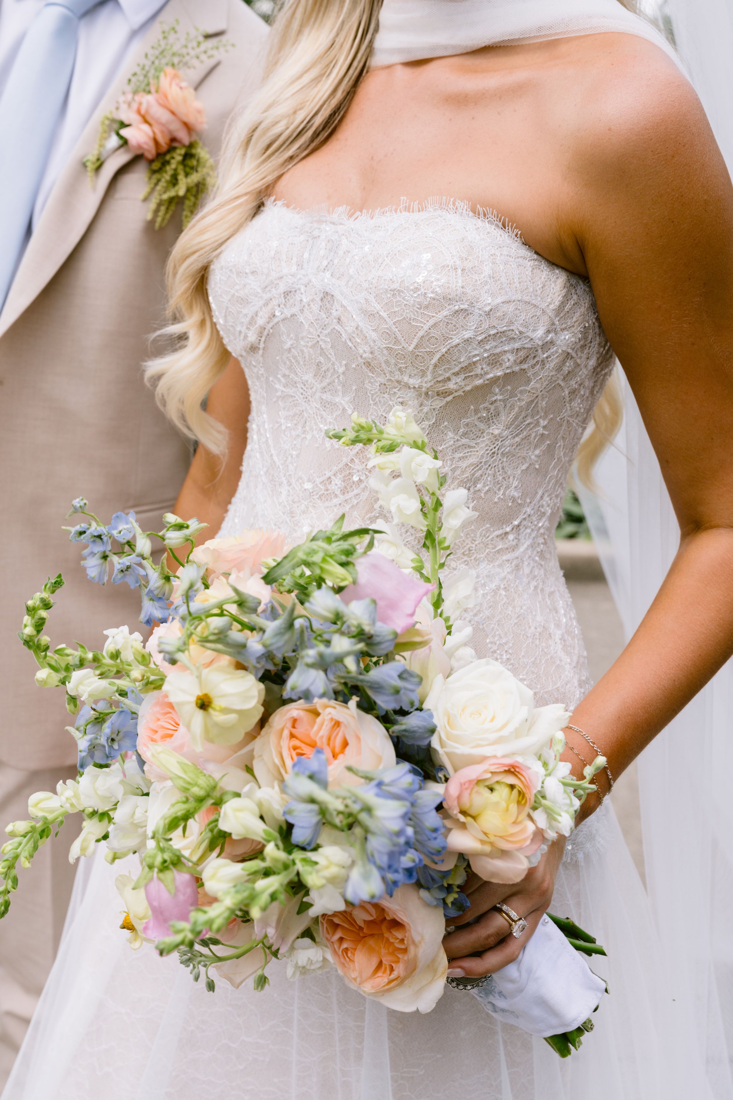 Bride in strapless beaded gown holding pastel bouquet with peach, white, and blue flowers beside groom in tan suit.
