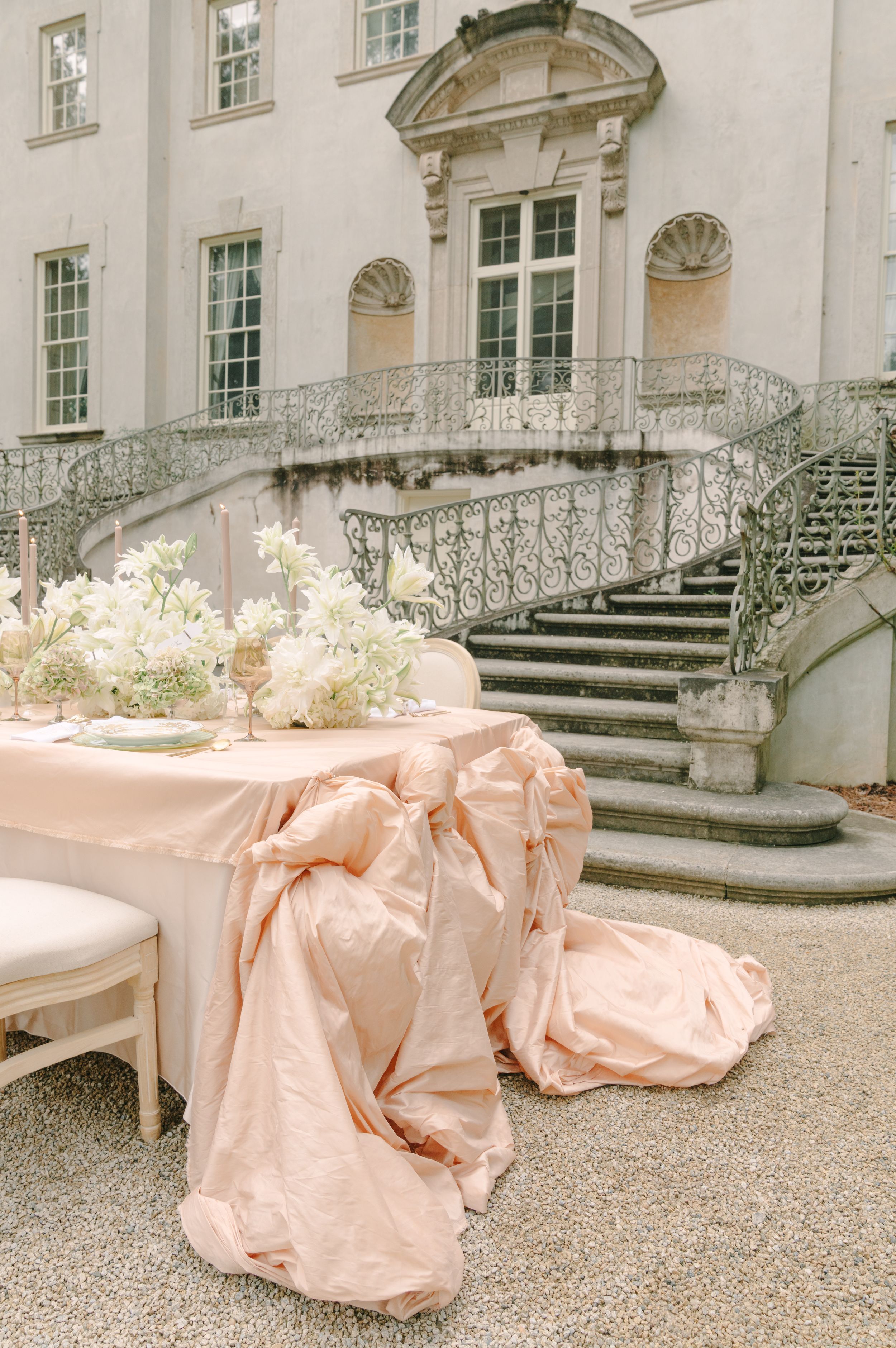 Elegant outdoor wedding table with blush pink linen and white floral centerpieces beside a grand mansion staircase.