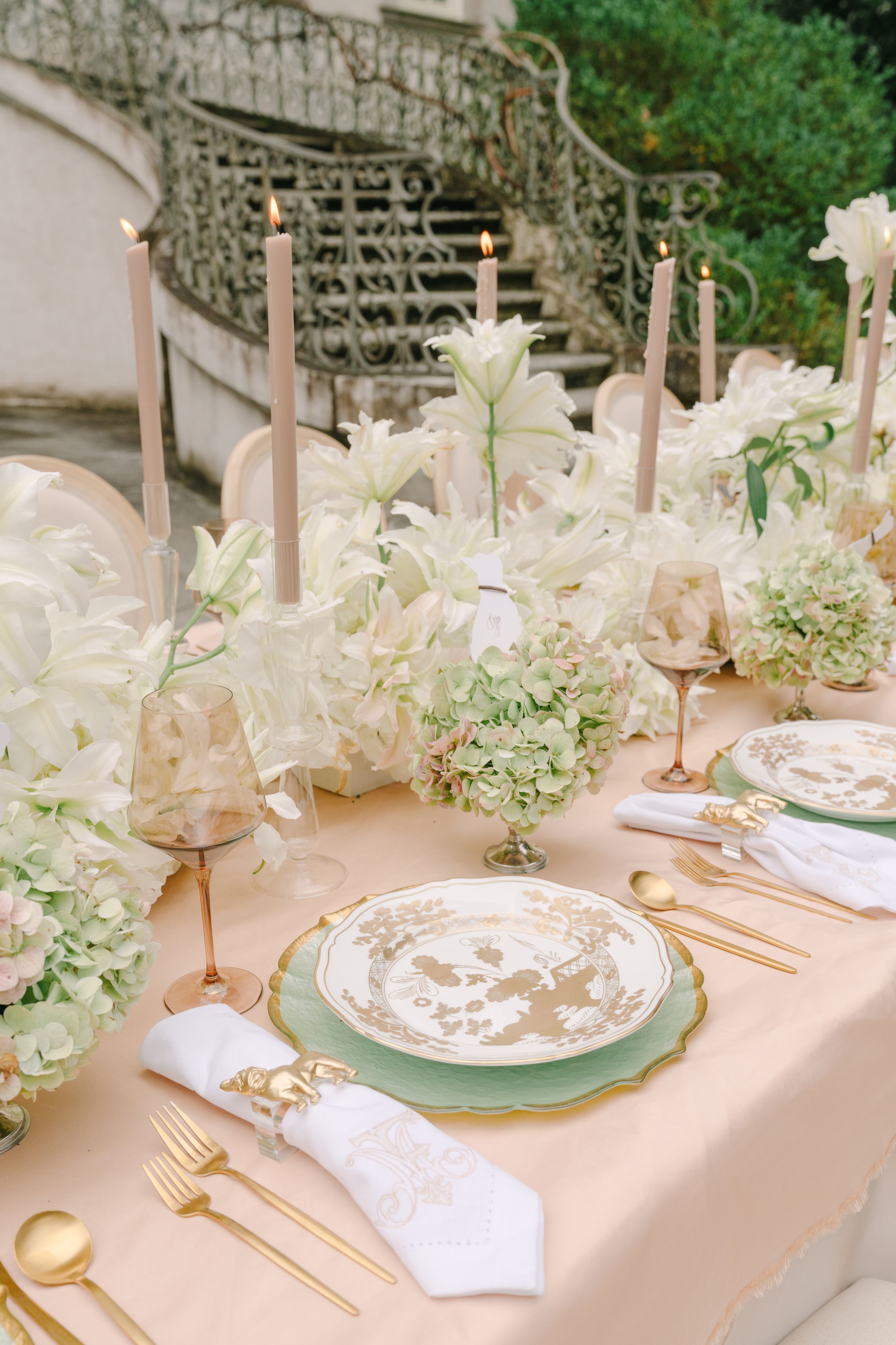 Elegant outdoor wedding table with gold cutlery, sage green plates, white floral arrangements, and blush candles on a peach tablecloth.