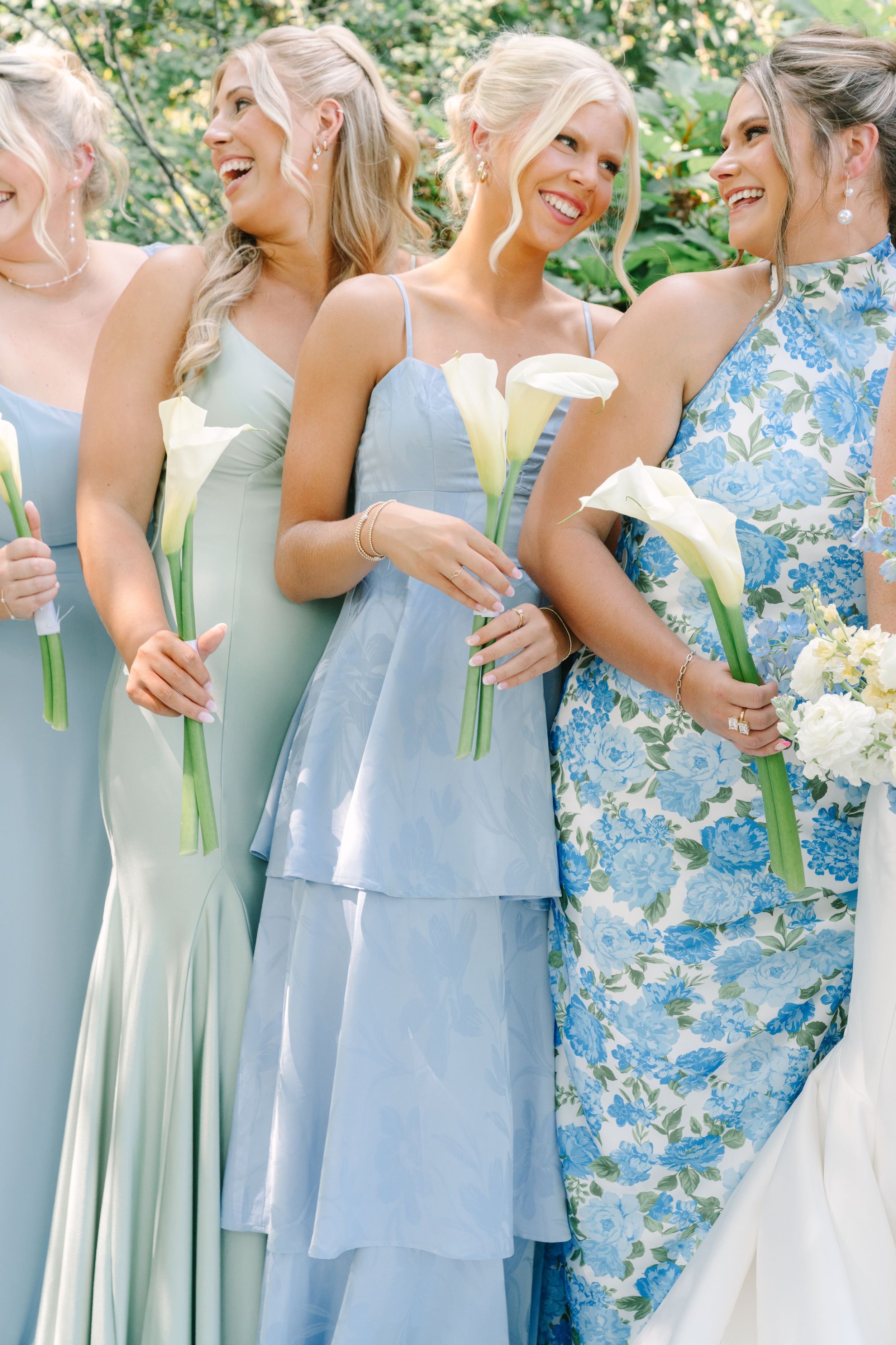 Bridesmaids in mismatched blue and sage green gowns holding white calla lilies, smiling outdoors at a wedding.