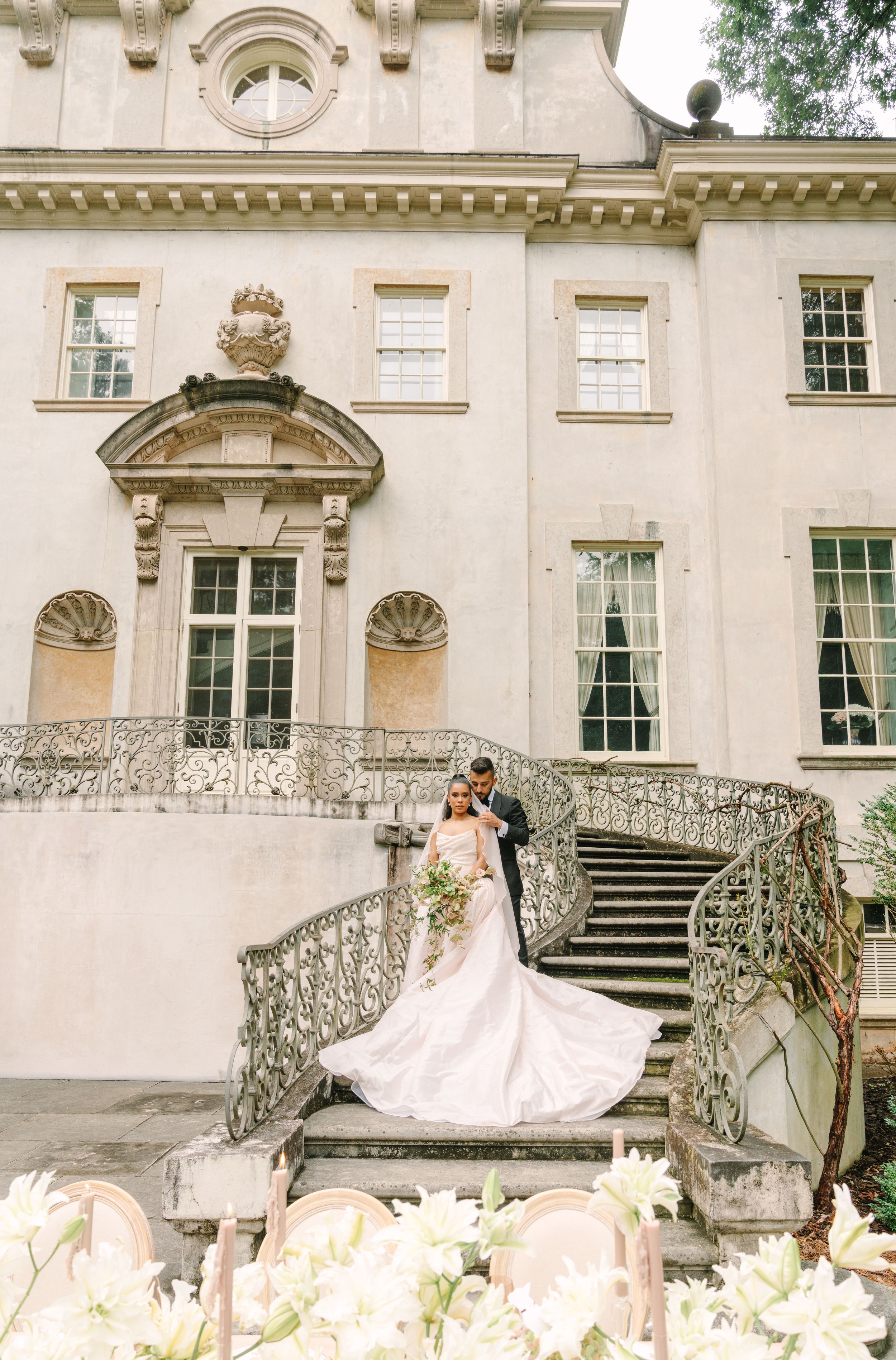 Bride and groom pose on ornate iron staircase of elegant European-style mansion in flowing wedding gown.