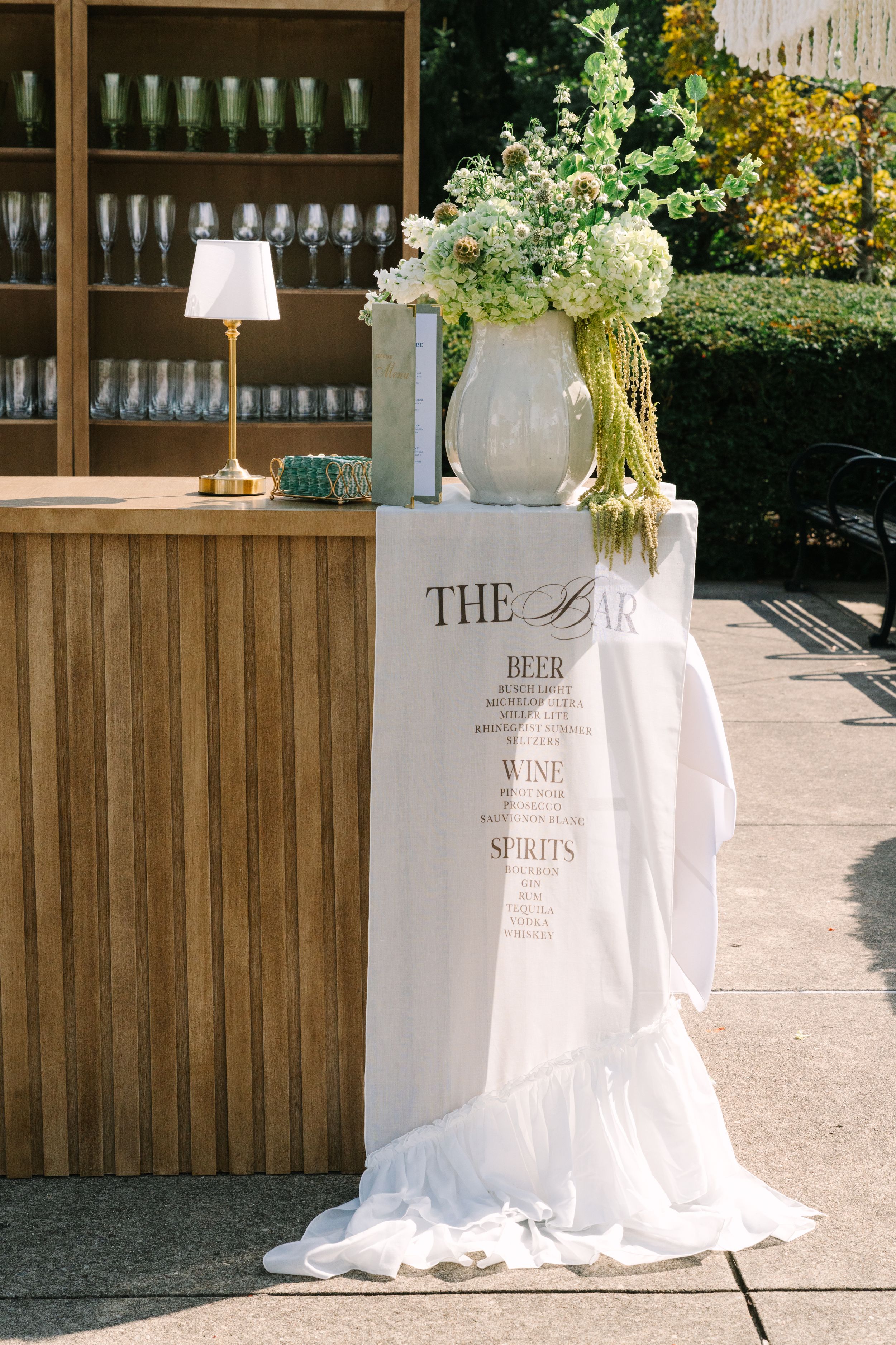 Elegant outdoor wedding bar setup with white floral arrangement, draped fabric menu sign listing beer, wine, and spirits.