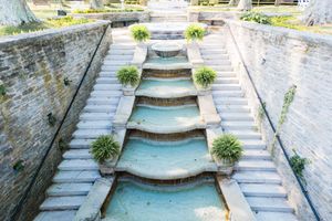 Ornate stone stepped fountain with cascading water levels and potted plants in a walled garden.