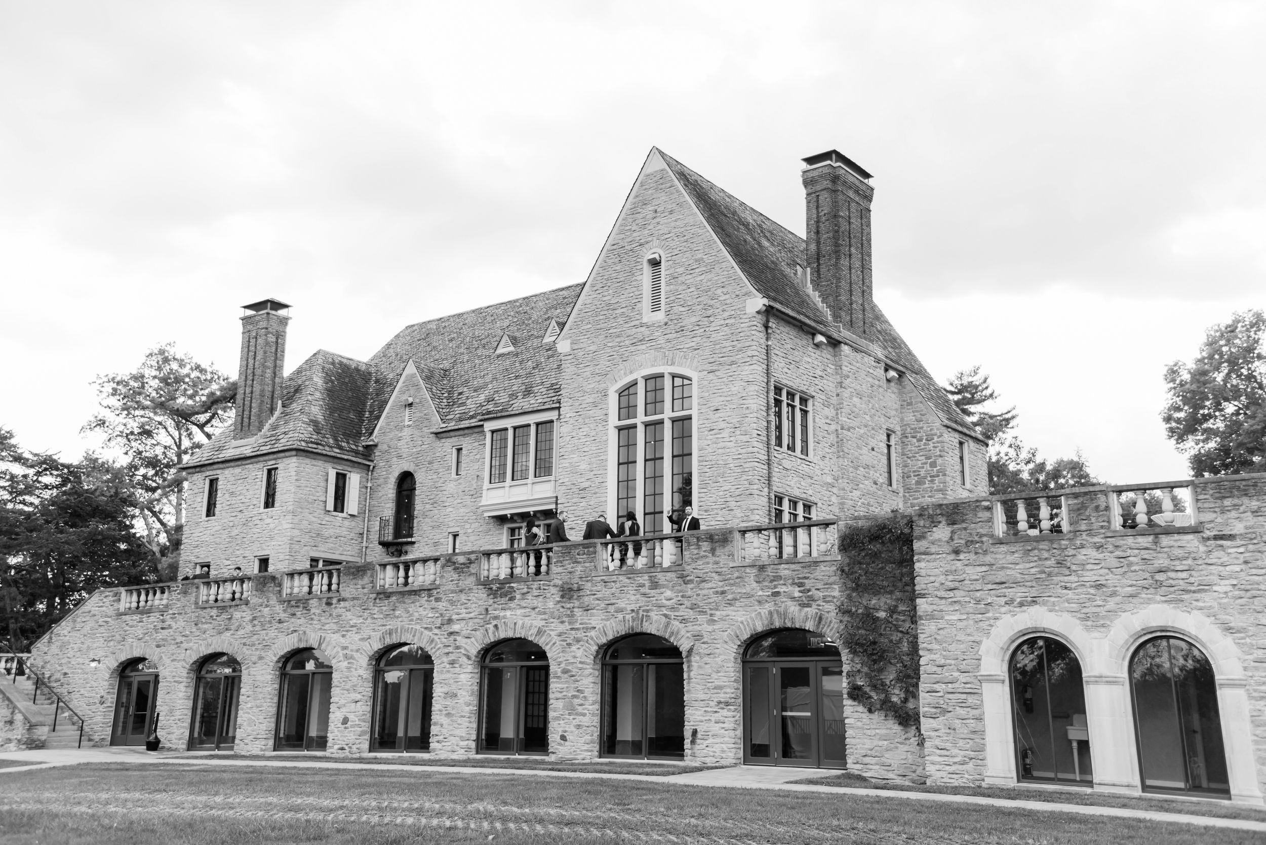 Black and white photo of an elegant stone manor house with arched windows, chimneys, and guests on the upper terrace.