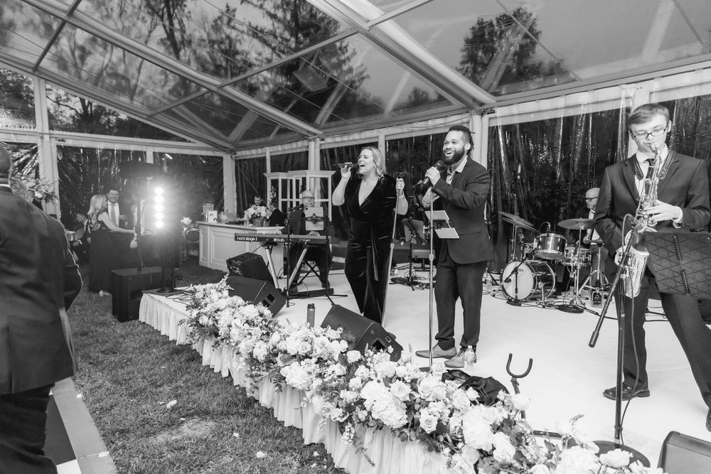 Live band performing at an elegant wedding reception under a clear tent with floral centerpieces.