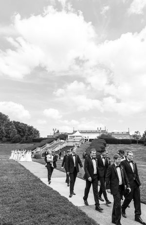 A wedding party in formal attire walks along a grassy hillside path under cloudy skies in black and white.