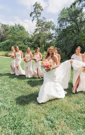 A bridal party in white dresses runs playfully through a grassy hillside on a sunny summer day.