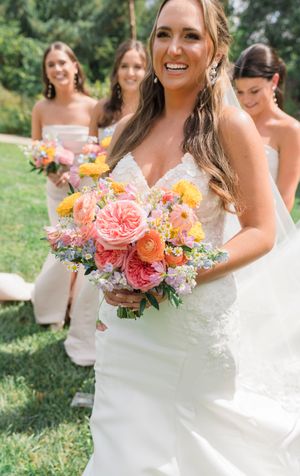 A summer wedding party poses outdoors with vibrant coral and yellow floral bouquets.
