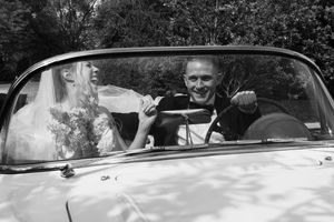 Black and white photo of a bride and groom smiling in a vintage convertible car on their wedding day.