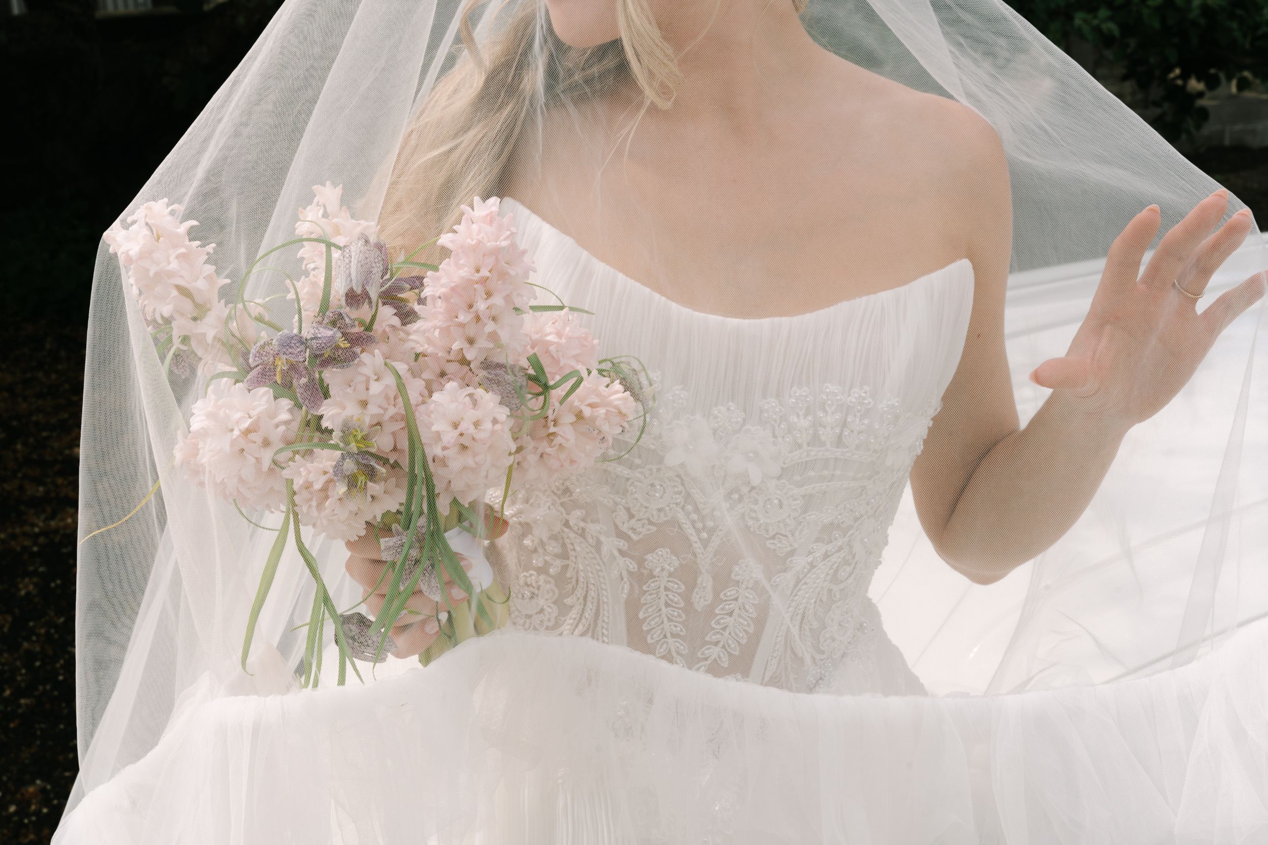 Bride in lace wedding gown holding soft pink floral bouquet under sheer veil in elegant bridal portrait.
