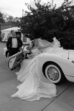 Black and white photo of bride in flowing gown exiting vintage white Corvette as groom in tuxedo stands nearby.