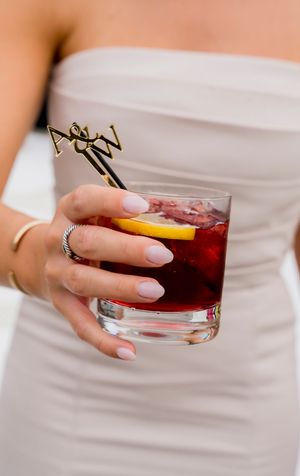 Hands holding red cocktails in crystal glasses against a white dress background.
