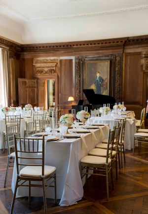 Elegant banquet hall setup with white-clothed round tables, chiavari chairs, and wood-paneled walls with classic artwork.