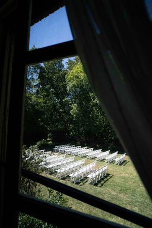 View through a window of white ceremony chairs set up on a lawn for an outdoor wedding.