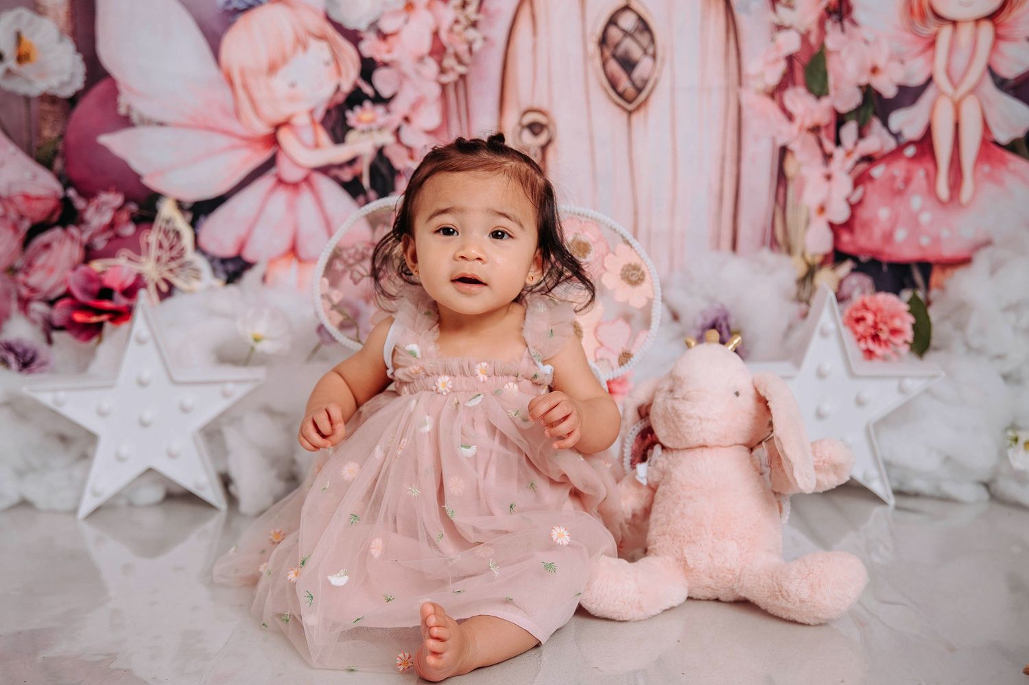 A baby in a pink tulle dress sits on a white floor surrounded by pink decorations and star accents.