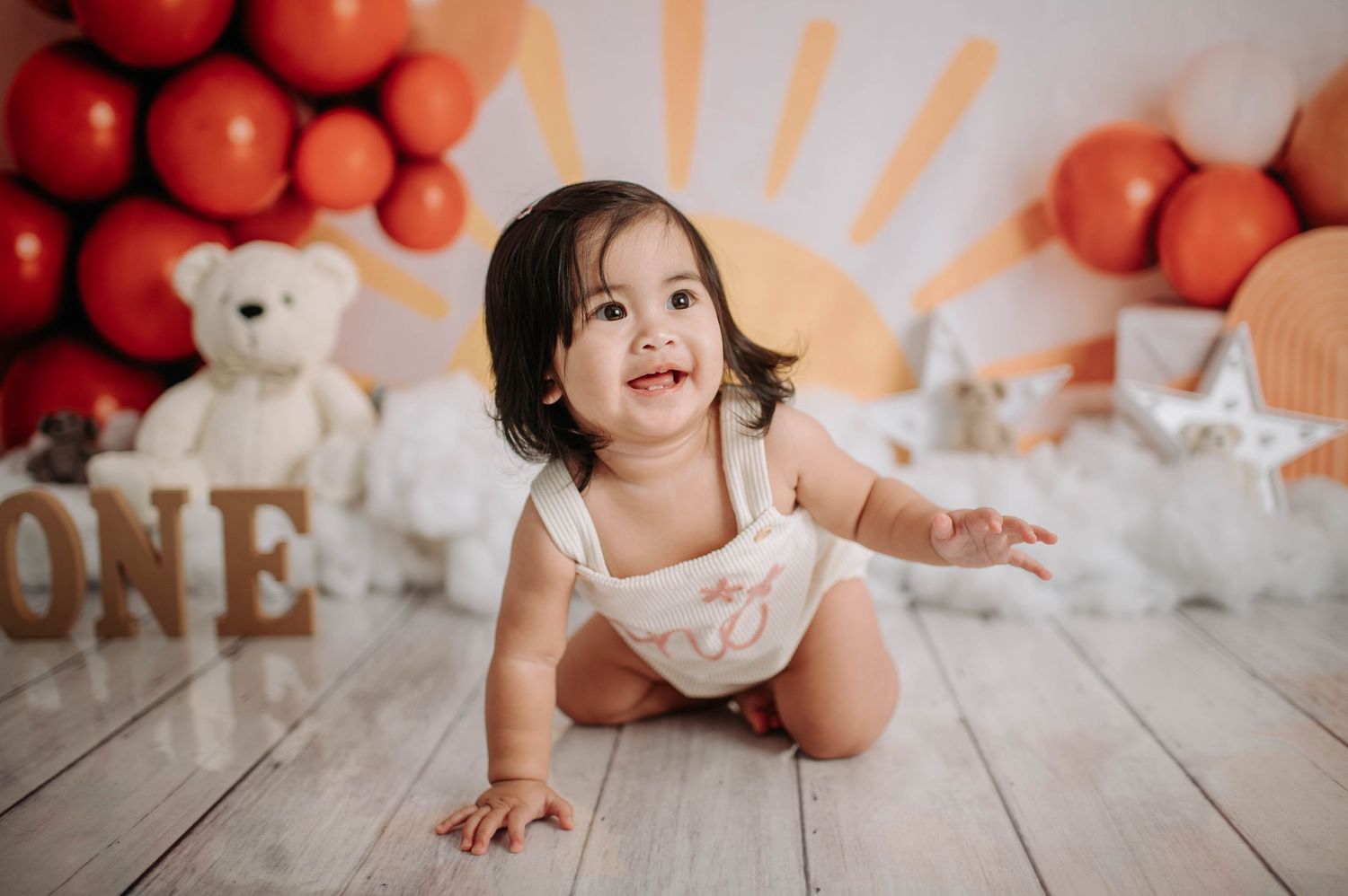 First birthday photoshoot setup with red balloon arch, teddy bear and 'ONE' text on white wooden floor.
