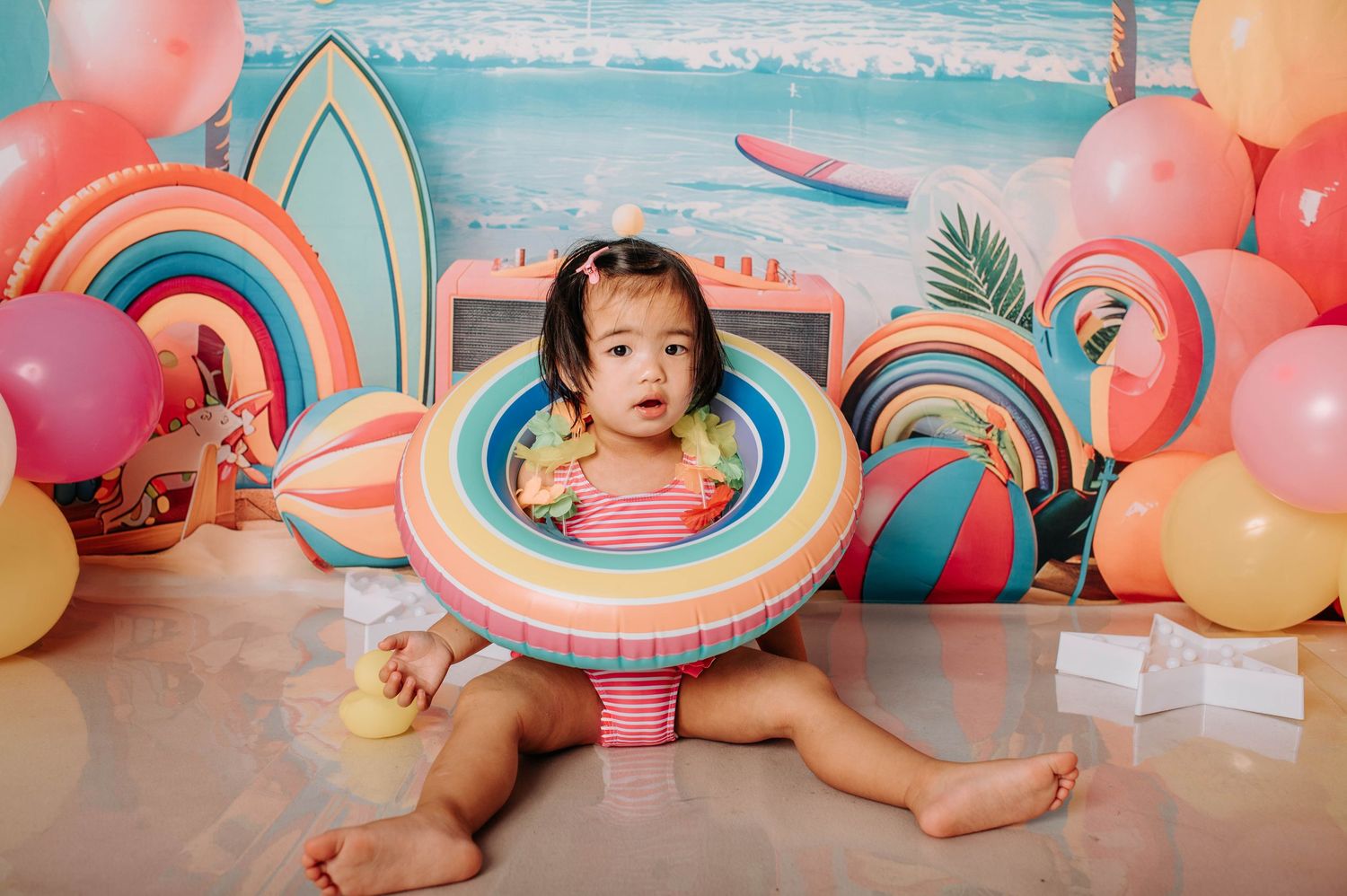 Colorful balloon and pool float decorations create a whimsical summer scene with pastel pink and blue tones.