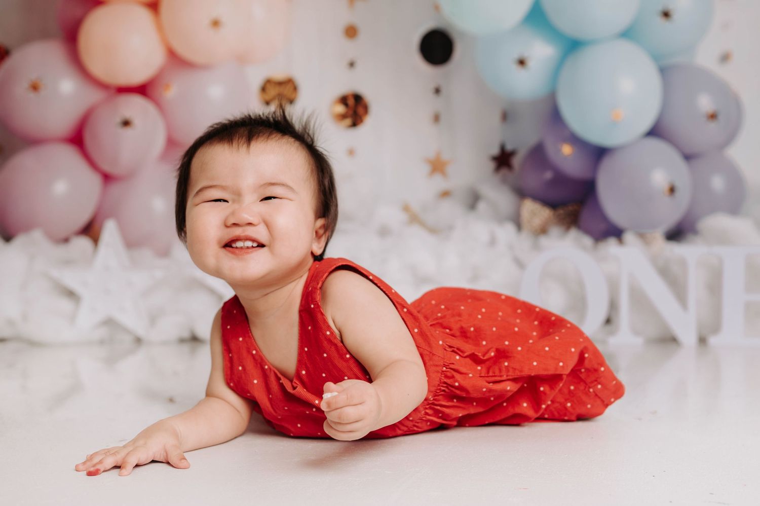 A baby wearing a red dress lies on a white floor with pastel balloon decorations and stars in the background.