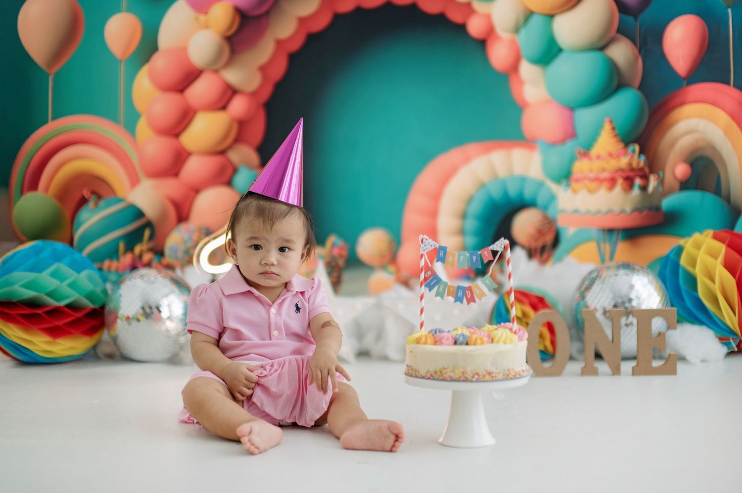Baby in pink dress celebrates first birthday with colorful balloon backdrop and smash cake setup.