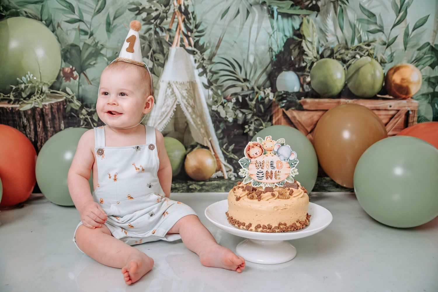 A cake smash photo session setup with sage green balloons, decorative greenery and a rustic birthday cake on a white stand.