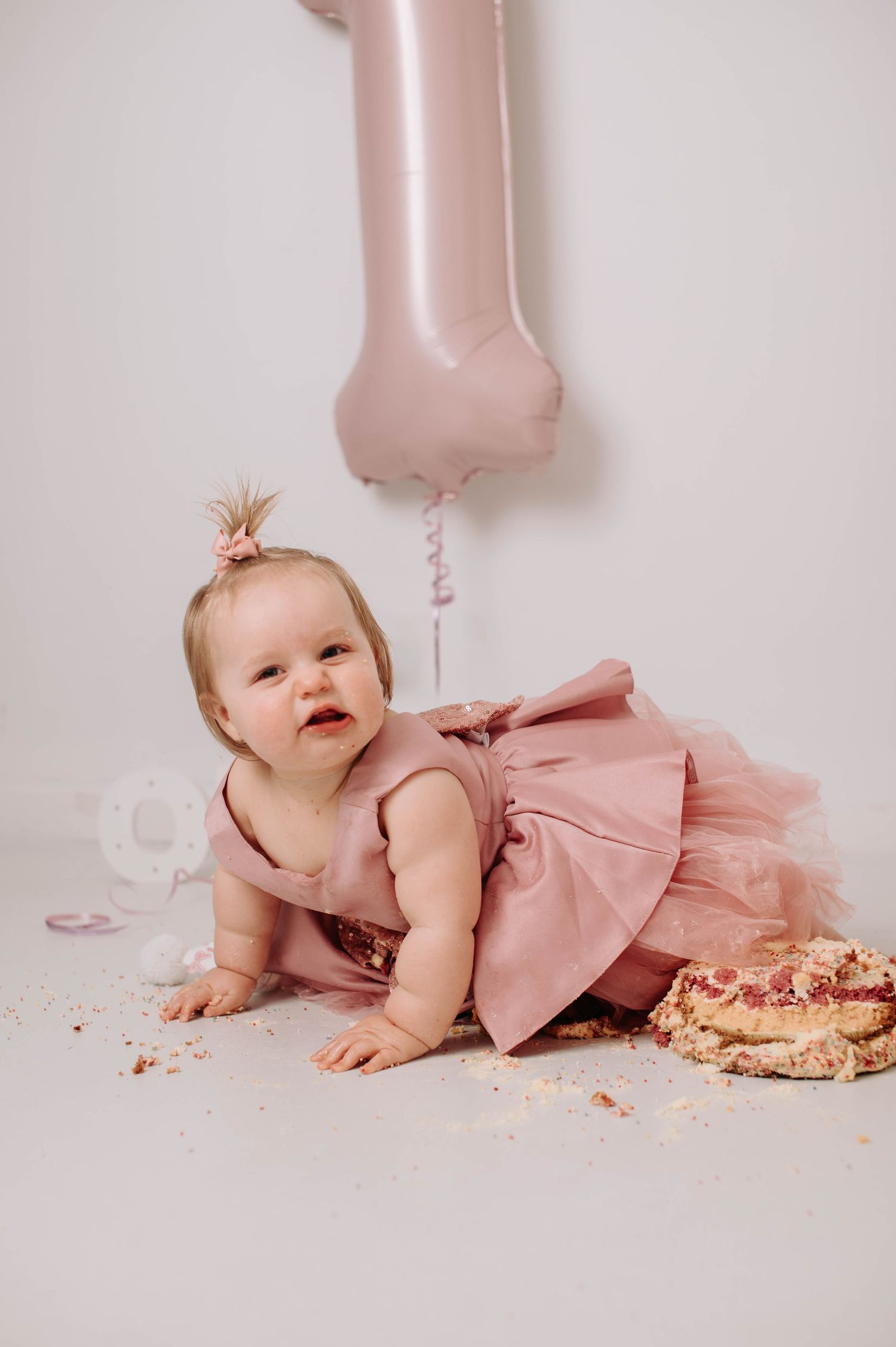 Baby in pink dress crawls and plays with smashed cake during first birthday photo session with rose gold balloon decor.
