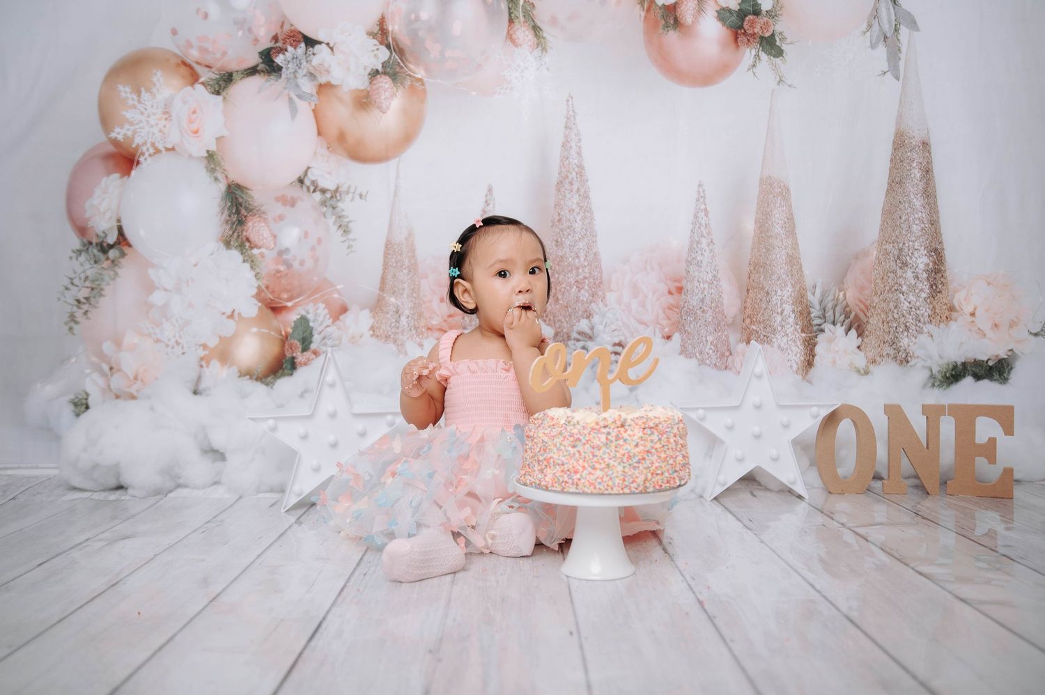 A pink and gold first birthday cake smash photo session setup with floral decorations and rustic woodland elements.