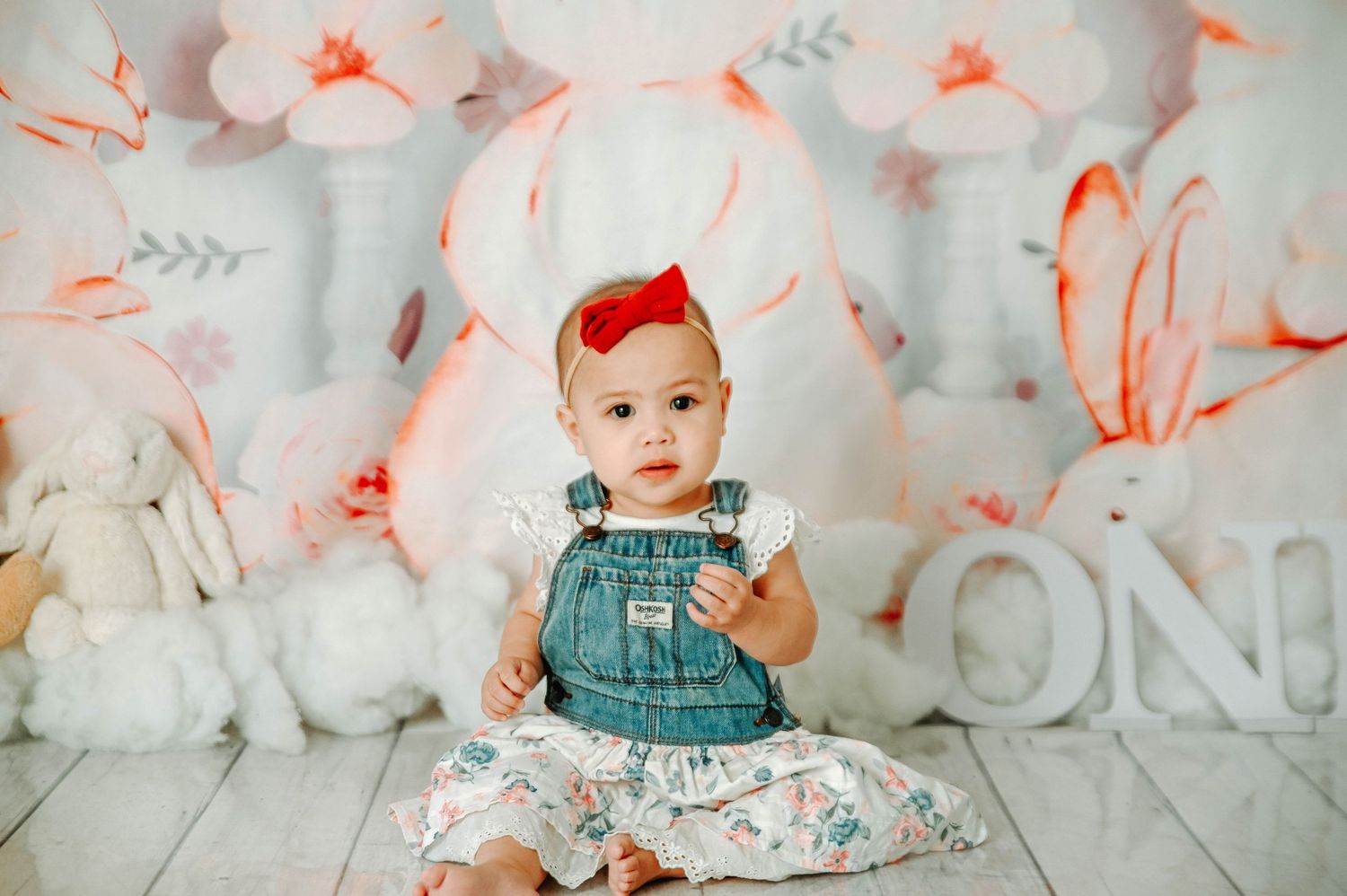 Baby in denim overalls sits against floral backdrop with coral and white paper flowers.