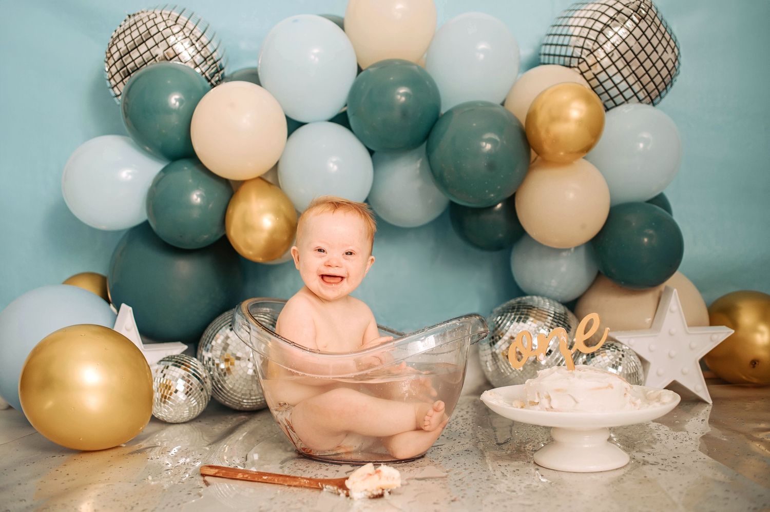Festive first birthday setup with sage green and gold balloons, white cake stand, and decorative elements on gray backdrop.