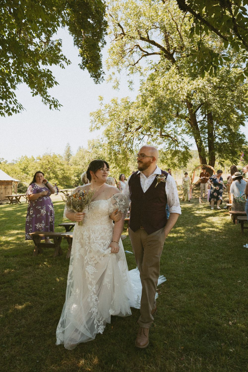 A couple dances together at an outdoor wedding reception under shady trees on a sunny afternoon.
