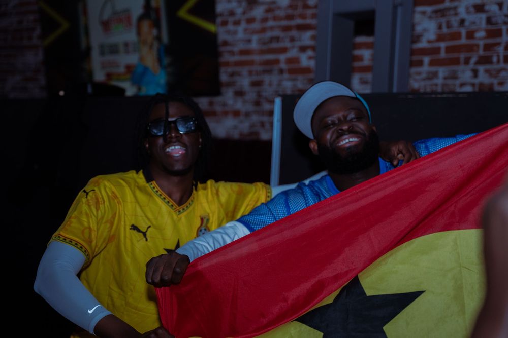 Two men smiling and holding a Ghana flag, wearing yellow jerseys, cheering at a sports viewing event.